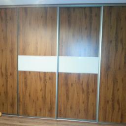 Meble Pod Wymiar - Czterodrzwiowa szafa wnękowa z przesuwnymi drzwiami, wykończona okleiną drewnopodobną w ciepłym odcieniu brązu, z poziomym pasem białego szkła na środku.