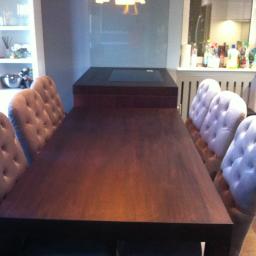 Meble Pod Wymiar - Ciemny, drewniany stół jadalny z sześcioma krzesłami tapicerowanymi w stylu Chesterfield, widoczne elementy wyposażenia kuchni w tle.