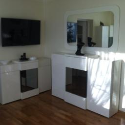 Meble Pod Wymiar - Nowoczesne, białe meble do salonu z elementami szklanymi, telewizor na ścianie, lustro z zaokrąglonymi rogami, drewniana podłoga.