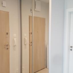 Meble Pod Wymiar - Szafa wnękowa z lustrzanymi drzwiami przesuwnymi w przedpokoju, odbijająca drzwi wejściowe z folią ochronną i biały domofon na ścianie.