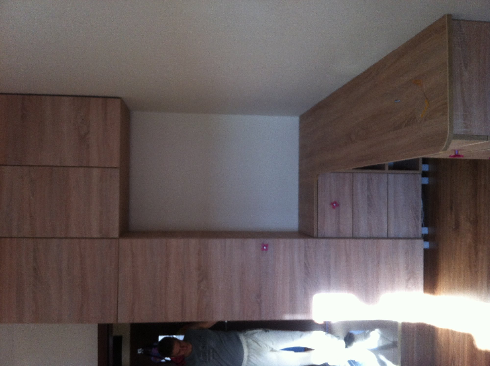 Zabudowa meblowa z drewnopodobnej płyty, widoczny fragment sufitu i odbicie osoby w błyszczącej powierzchni