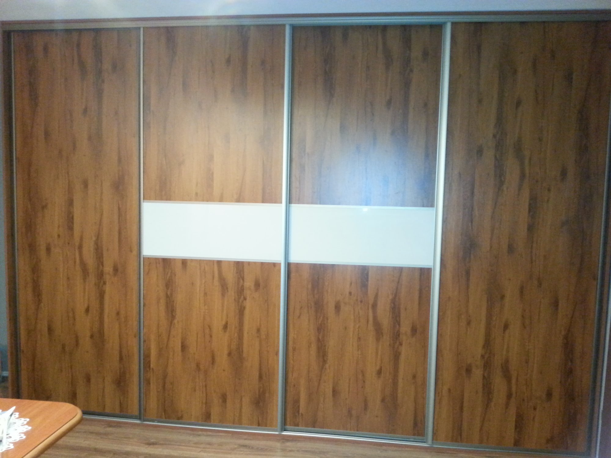 Czterodrzwiowa szafa wnękowa z przesuwnymi drzwiami, wykończona okleiną drewnopodobną w ciepłym odcieniu brązu, z poziomym pasem białego szkła na środku.