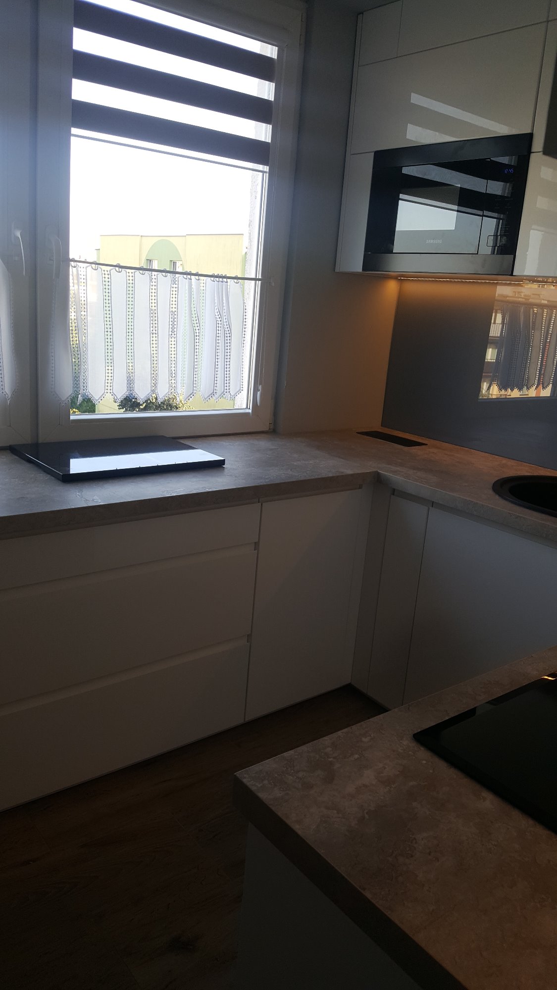 Nowoczesna, jasna kuchnia z białymi szafkami, blatem imitującym kamień i oknem z roletami dzień-noc oraz firanką z haftem w tle, widoczny fragment płyty indukcyjnej i zabudowany piekarnik.