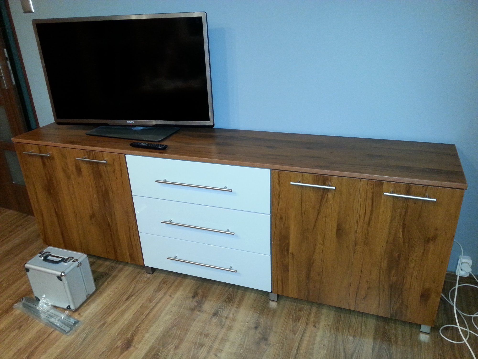 Szafka RTV z drewnopodobnymi frontami i trzema białymi szufladami, na blacie telewizor, obok skrzynka z okuciami do montażu szuflad.