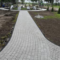 F.P.H.U Dan-Bud - Ścieżka z jasnoszarej kostki ułożona w łukowaty wzór, otoczona rabatami z młodymi drzewkami i ozdobnymi kamieniami, z pracującym robotnikiem i traktorem w tle.