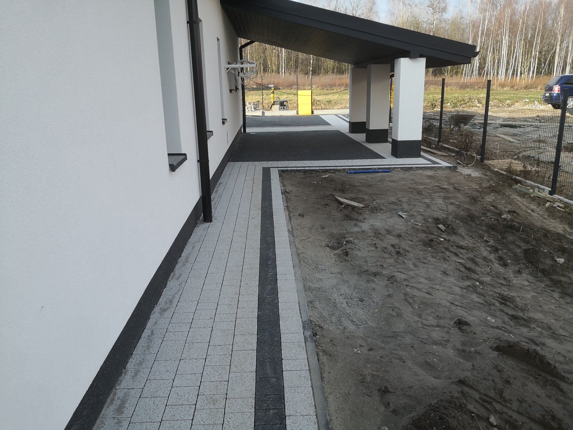 Nowoczesny podjazd z jasnoszarej kostki brukowej z ciemnoszarym pasem, prowadzący do zadaszonego wejścia domu z białymi kolumnami, widok na przygotowany teren pod ogród.