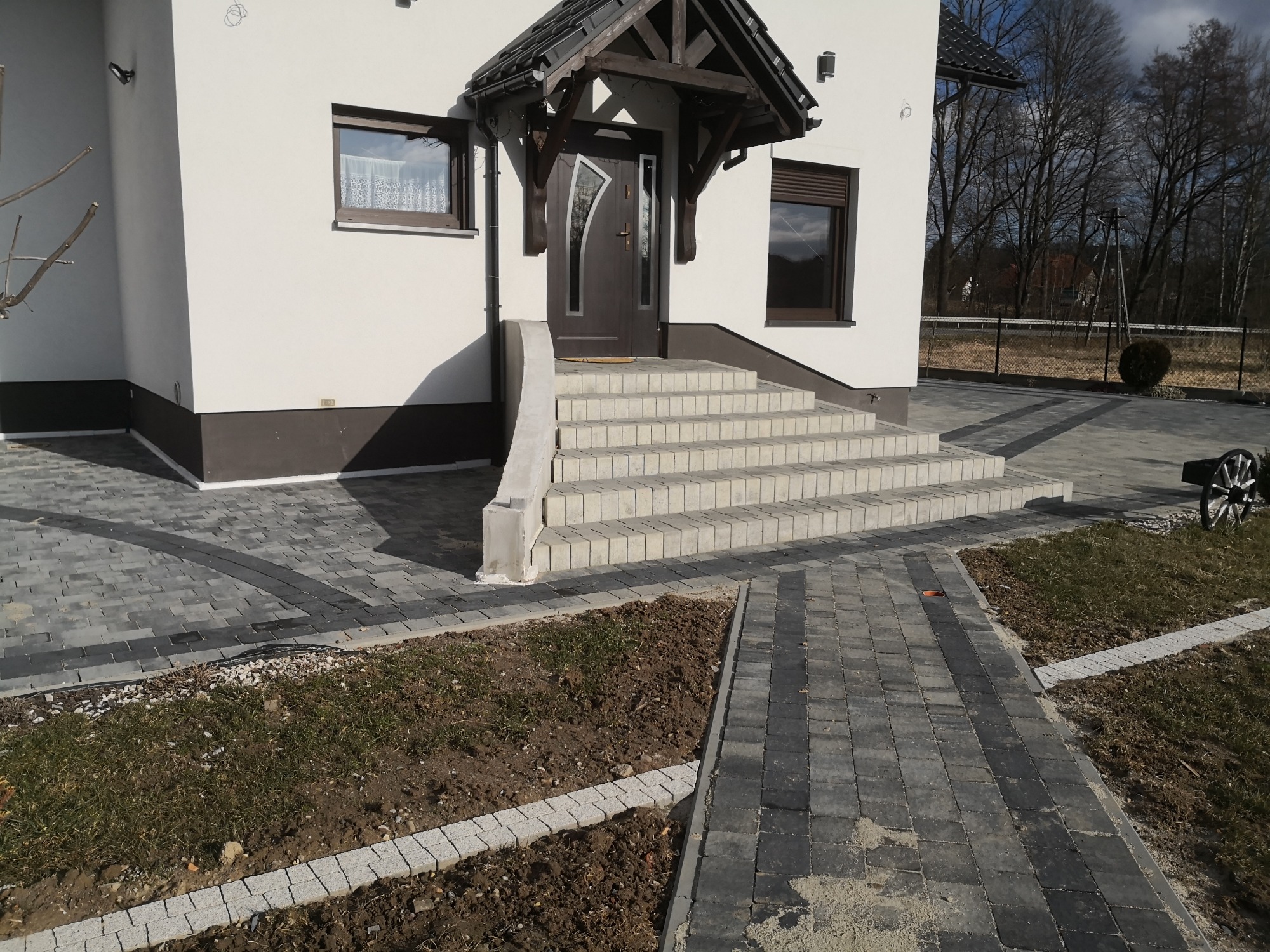 Ułożona kostka brukowa w dwóch odcieniach szarości prowadząca do wejścia domu z drewnianą wiatą, widoczne schody z kostki i obrzeża.