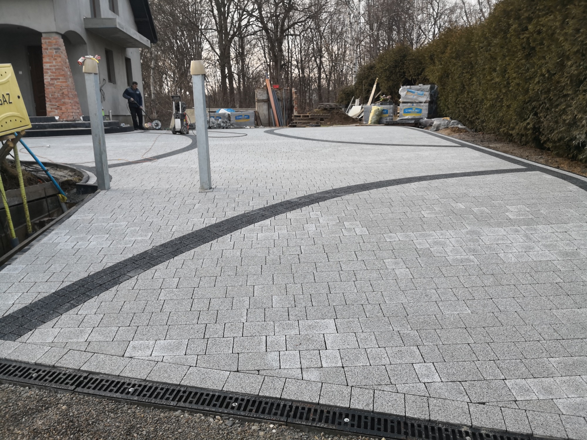 Ułożona szara kostka brukowa z dekoracyjnymi, ciemnymi pasami, widoczny pracownik z myjką ciśnieniową i przygotowane palety kostki w tle.