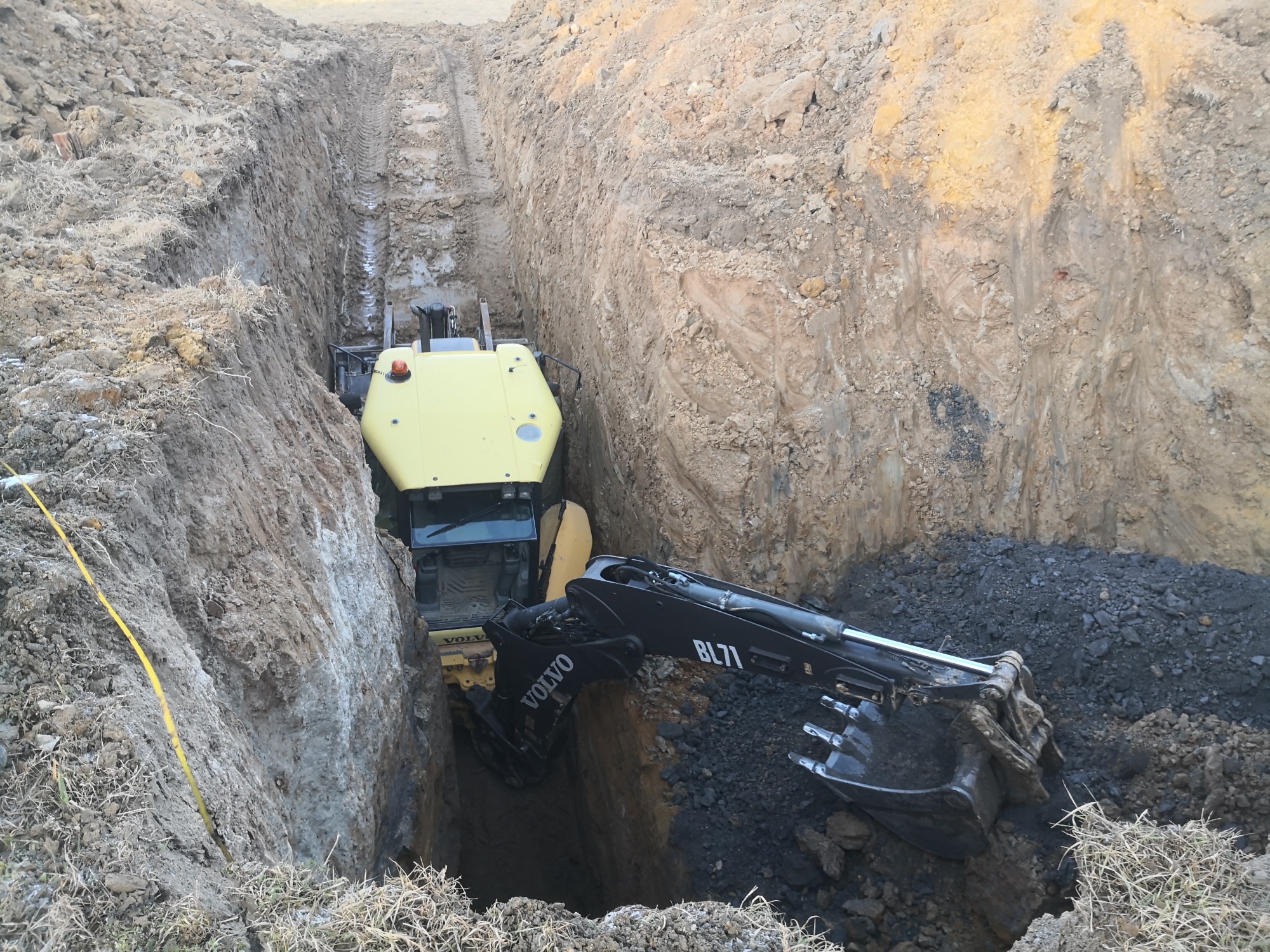 Żółto-czarna koparka Volvo BL71 podczas prac ziemnych w głębokim wykopie z widocznymi śladami opon na ścianach.