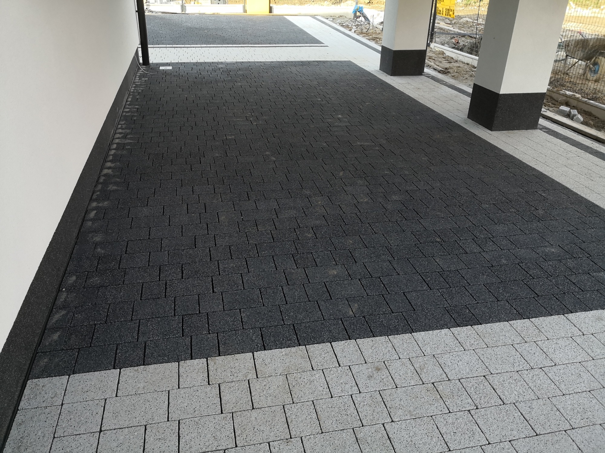 Ułożona nawierzchnia z kostki brukowej w odcieniach szarości, z obramowaniem z jasnych kostek, przylegająca do białej ściany budynku i filarów, widoczne prace wykończeniowe w tle.