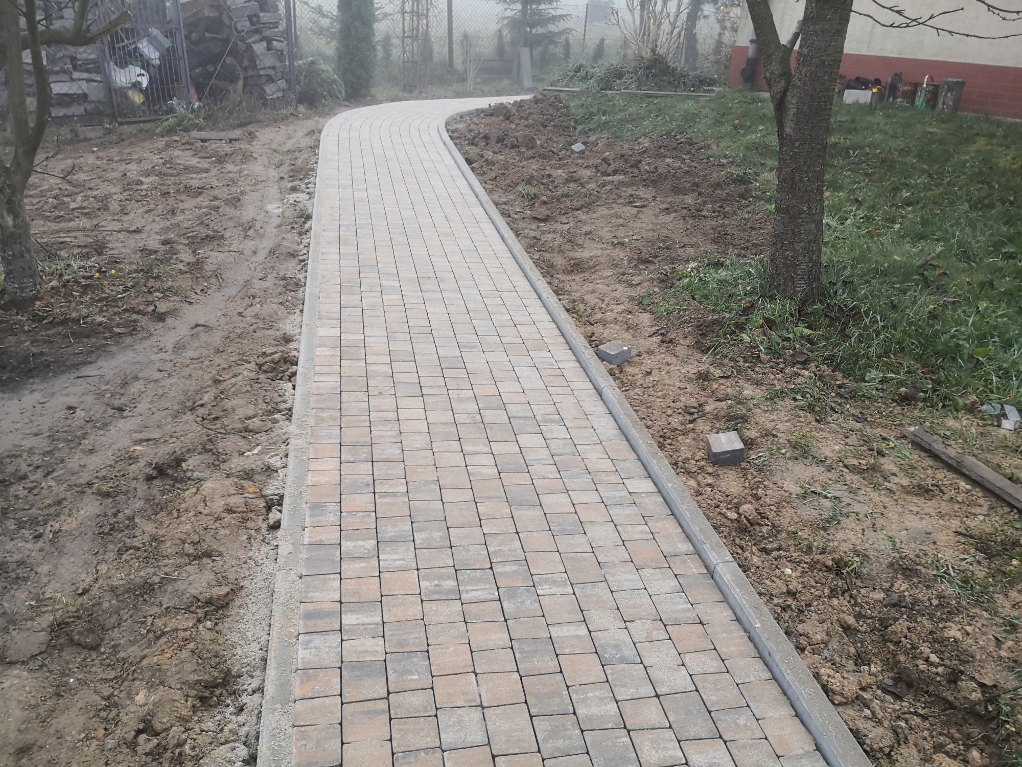 Zakręcający ścieżką ułożona z kostki brukowej w odcieniach szarości i beżu, z widocznymi elementami krajobrazu w pochmurny dzień.