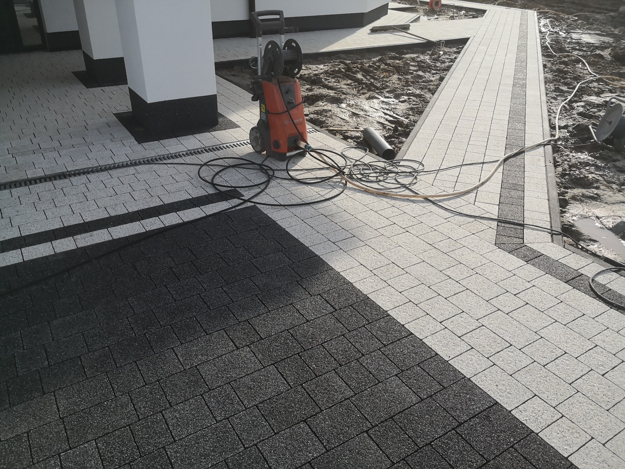 Ułożona dwukolorowa kostka brukowa przed budynkiem, z widocznym odpływem liniowym, myjką ciśnieniową i fragmentem terenu przygotowanego pod dalsze prace.
