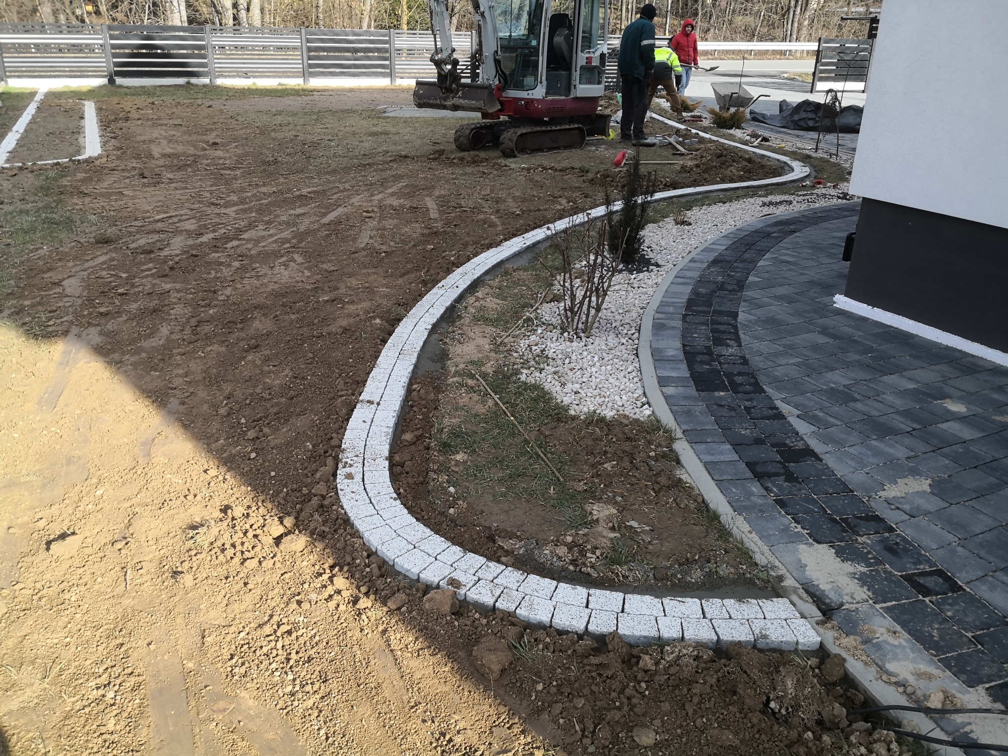 Budowa ścieżki z kostki granitowej i obrzeży z białej kostki, widoczny mini-koparka i pracownicy w tle, teren przygotowany pod trawnik.