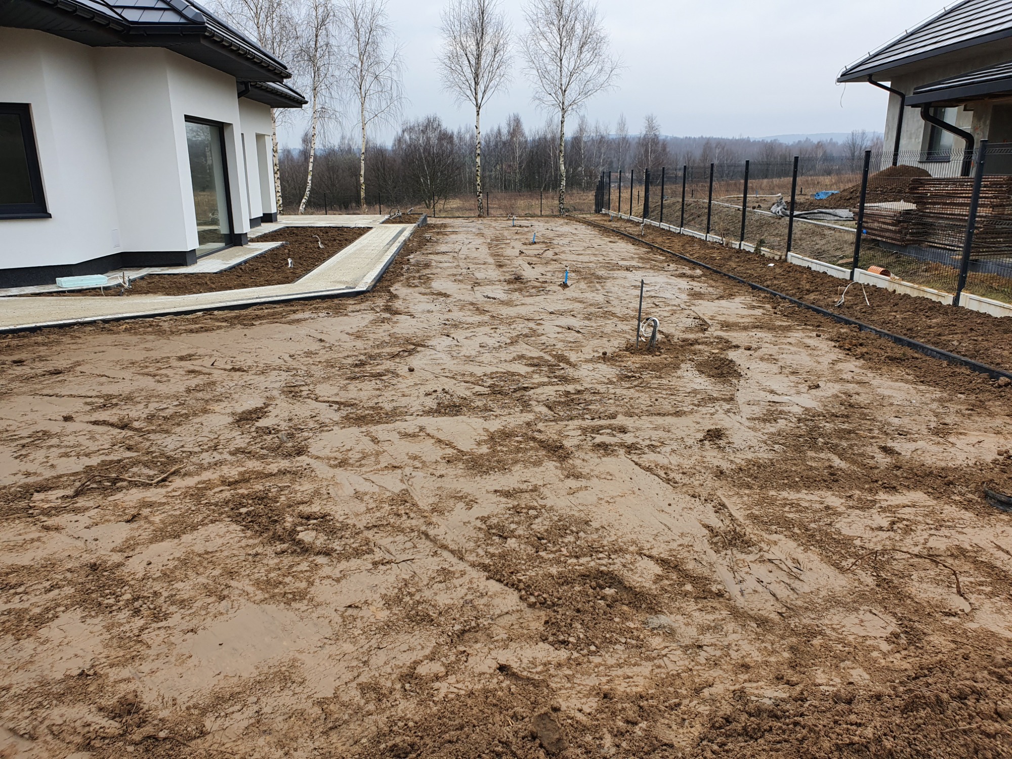 Przygotowany teren pod budowę z częściowo ułożoną kostką brukową wokół domu, widoczne wystające rury instalacyjne i nowa metalowa siatka ogrodzeniowa w tle.