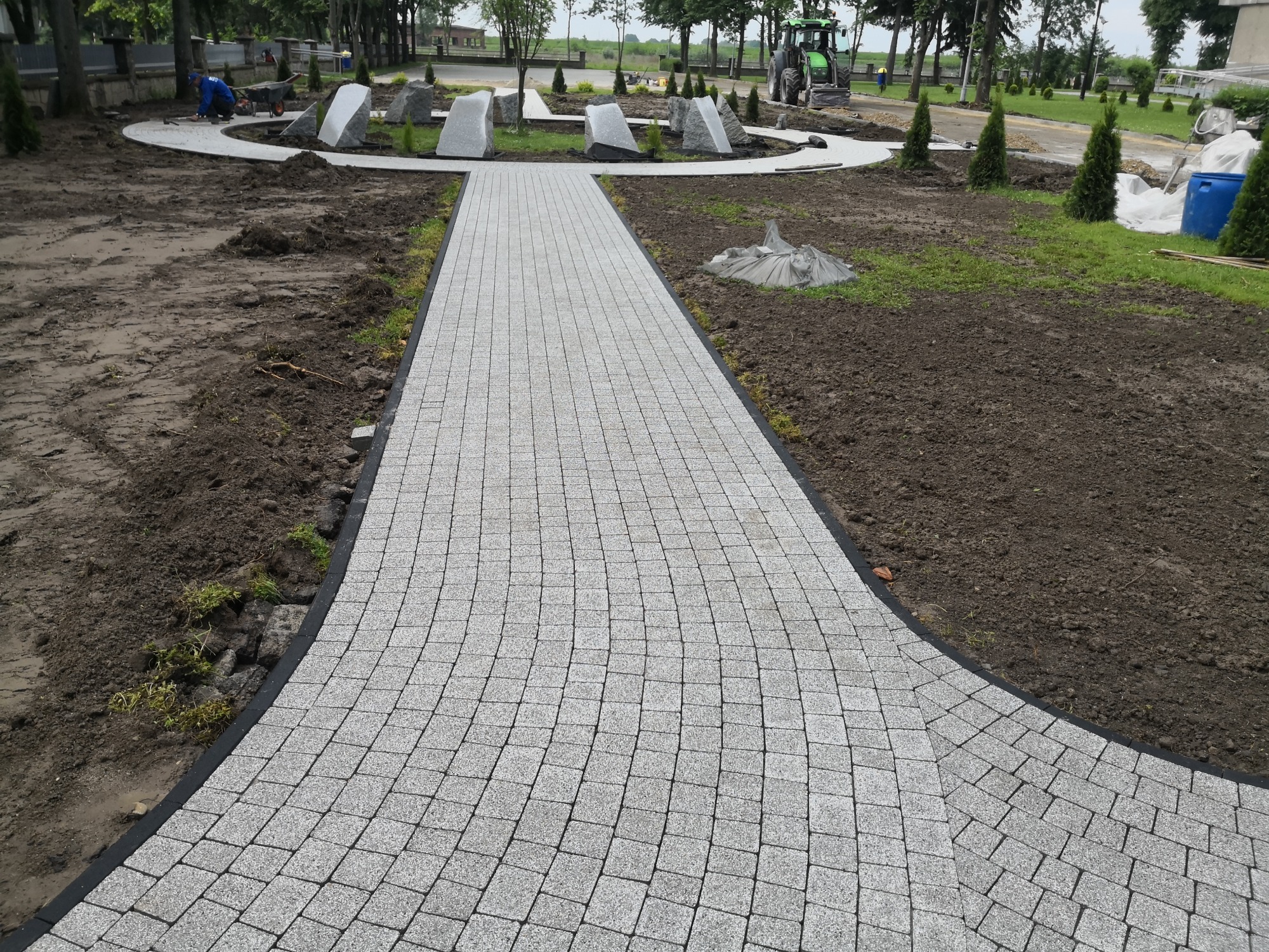 Ścieżka z jasnoszarej kostki ułożona w łukowaty wzór, otoczona rabatami z młodymi drzewkami i ozdobnymi kamieniami, z pracującym robotnikiem i traktorem w tle.