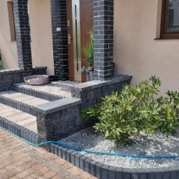 Schody wejściowe do domu wyłożone szarą kostką brukową, obrzeża z ciemnej kostki, rabata z białym grysem i zielonymi krzewami, niebieski wąż ogrodowy wzdłuż obrzeża.