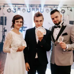 Iluzjonista prezentuje wachlarz kart, a para w strojach galowych trzyma pojedyncze karty na tle sceny z oświetleniem.