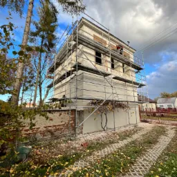 Dwupiętrowy budynek w trakcie prac elewacyjnych, otoczony rusztowaniem; w tle widoczne drzewa i błękitne niebo z chmurami; pracownik w czerwonej koszuli na rusztowaniu.