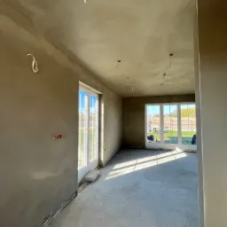 Wnętrze domu w trakcie prac wykończeniowych: ściany pokryte świeżym, surowym tynkiem maszynowym, widoczne wystające przewody elektryczne i osadzone puszki elektryczne, surowa betonowa wylewka...