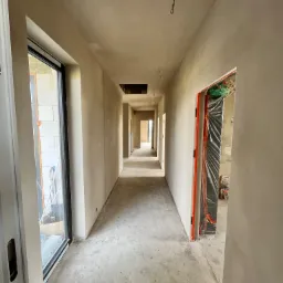 Długi korytarz w surowym stanie, ściany pokryte świeżym tynkiem, widoczne instalacje elektryczne na suficie, okno z widokiem na budowę, podłoga betonowa przed wylewką.