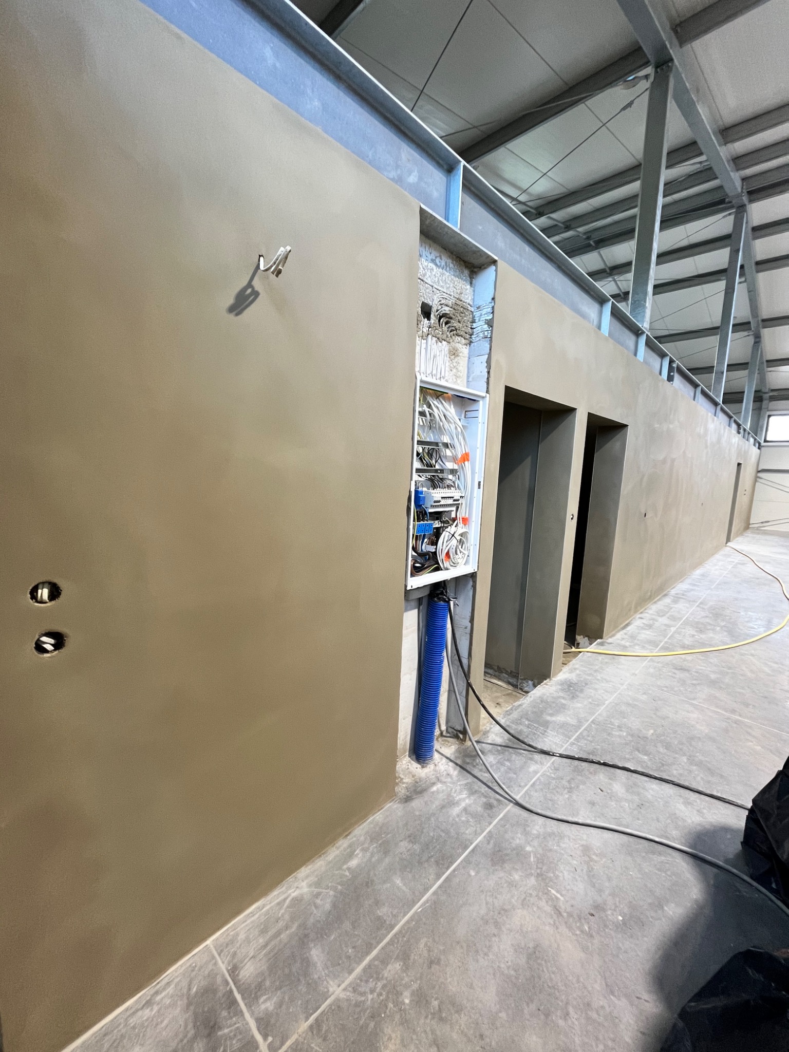Wnętrze hali przemysłowej w trakcie prac wykończeniowych, świeżo nałożone tynki na ścianach, widoczna instalacja elektryczna w otwartej skrzynce, metalowa konstrukcja dachu.