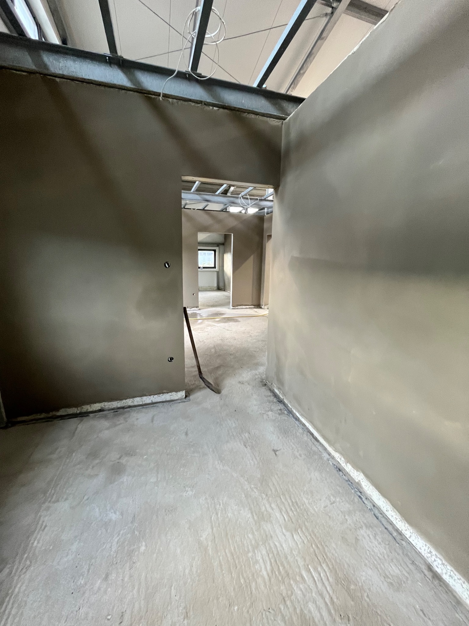 Wnętrze w trakcie tynkowania maszynowego, widoczne surowe ściany pokryte tynkiem, metalowa konstrukcja sufitu i łopata oparta o ścianę w przejściu.