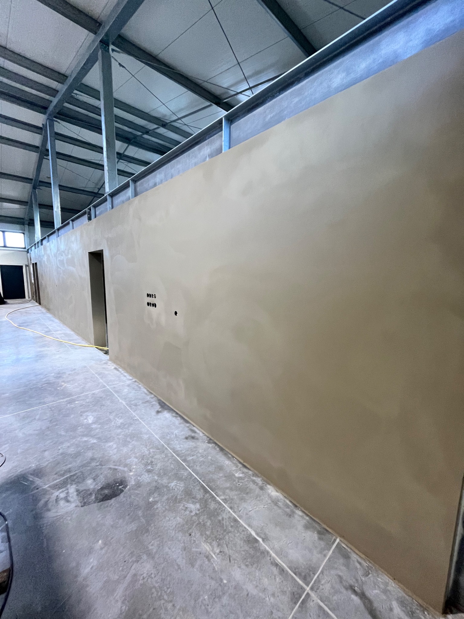 Świeżo nałożony, gładki tynk na ścianie w surowym wnętrzu hali, widoczne puszki instalacyjne, metalowa konstrukcja stropu i betonowa posadzka z fugami.