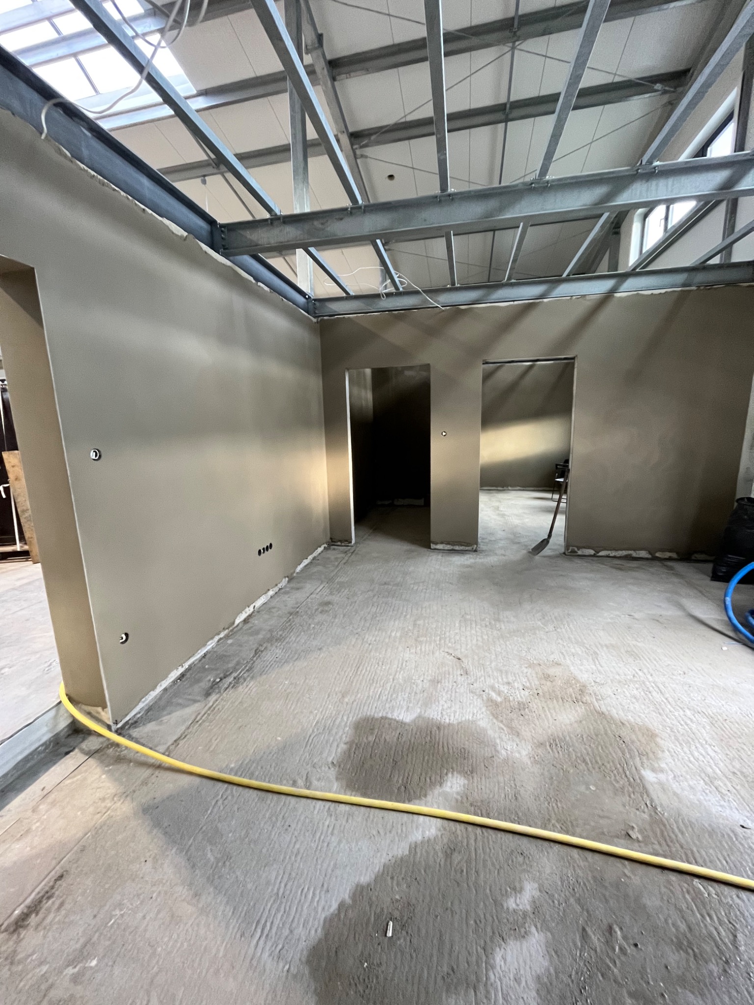 Wnętrze w trakcie prac wykończeniowych; świeżo położone gładzie na ścianach w odcieniach beżu, widoczne metalowe elementy konstrukcyjne pod sufitem, mokra posadzka, żółty kabel na podłodze i łopata...