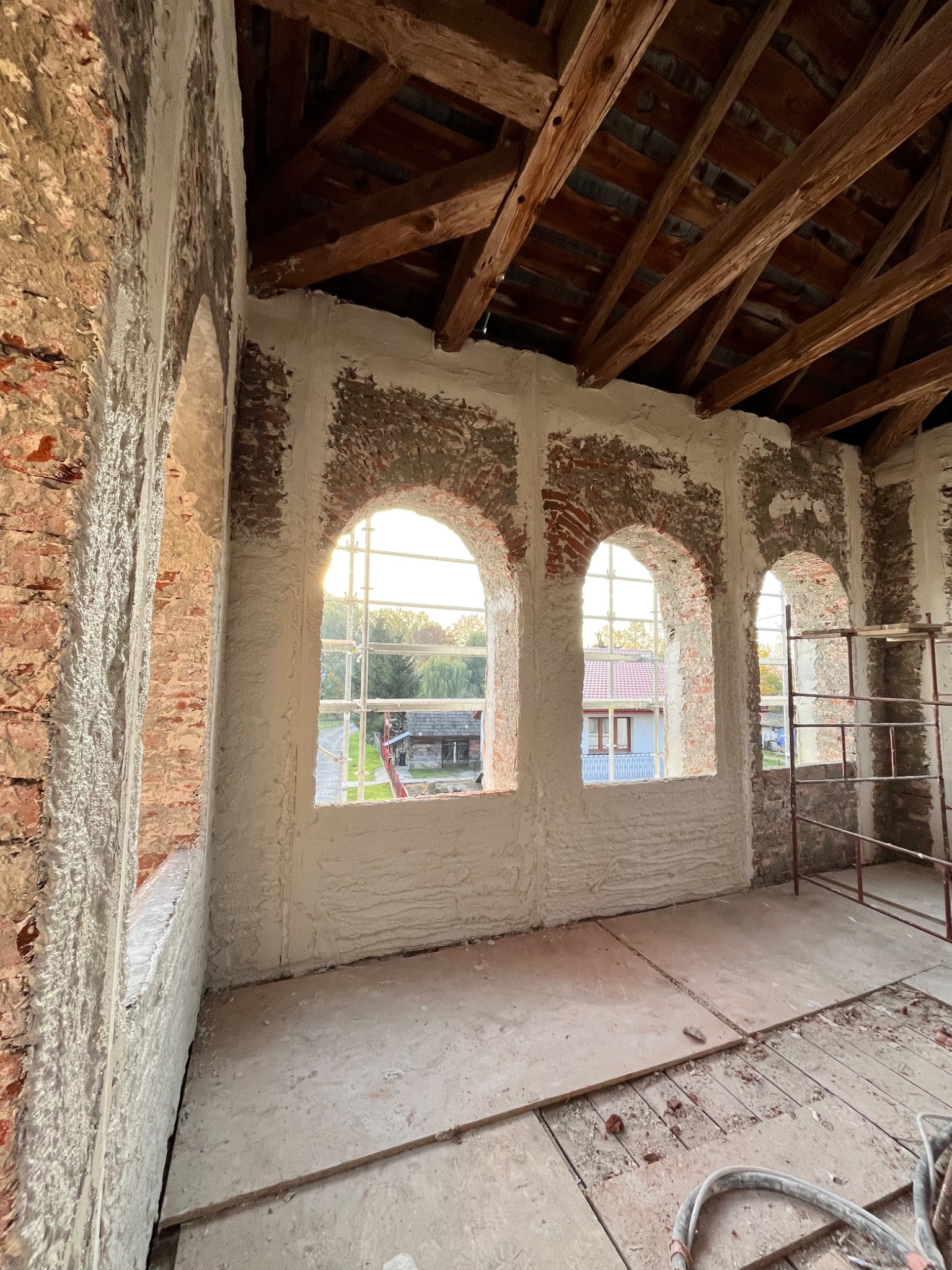 Wnętrze budynku w trakcie prac. Ściany z cegły częściowo pokryte białą warstwą, widoczne drewniane belki stropowe, okna łukowe z widokiem na zewnątrz, rusztowanie w rogu pomieszczenia.
