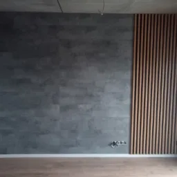 Ściana w Czempiń z szarymi panelami imitującymi beton i dekoracyjnymi drewnianymi lamelami po prawej stronie, widoczne gniazdka elektryczne.