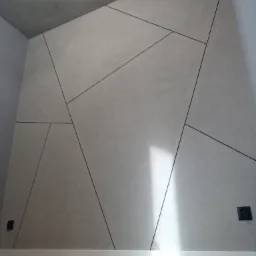 Dekoracyjna ściana w pokoju z geometrycznym wzorem wykonanym z czarnych linii na jasnoszarym tle, widoczne gniazdka elektryczne i fragment podłogi z paneli.