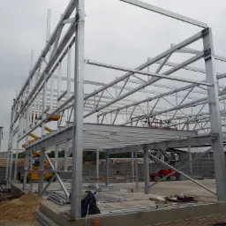 Stalowa konstrukcja hali w trakcie montażu, widoczny szkielet budynku, żółty podnośnik nożycowy i czerwony podest ruchomy, betonowa podstawa, pochmurne niebo w tle.