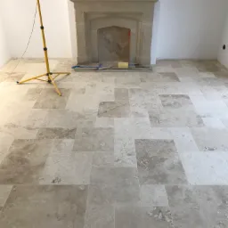 Ułożona podłoga z jasnych, naturalnych kamiennych płytek o nieregularnych kształtach, widoczny fragment kominka z kamienną obudową, ż&oacute;łta lampa robocza na tr&oacute;jnogu w rogu pomieszczenia.