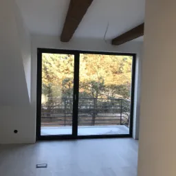 Wnętrze pokoju z drewnianymi belkami stropowymi, jasnymi panelami podłogowymi i dużym oknem balkonowym z widokiem na zewnątrz.