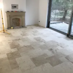 Wnętrze w trakcie prac wykończeniowych: ułożona podłoga z płytek, surowe ściany z przygotowanym miejscem na kominek, duże przesuwne okno z widokiem na zewnątrz, żółta lampa budowlana.