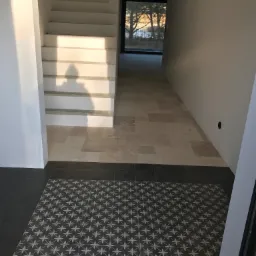 Wnętrze domu w trakcie wykańczania, z widocznymi schodami, dwiema rodzajami płytek podłogowych - wzorzystymi ciemnymi i beżowymi, oraz świeżo pomalowanymi ścianami w jasnych odcieniach. Na schodach...