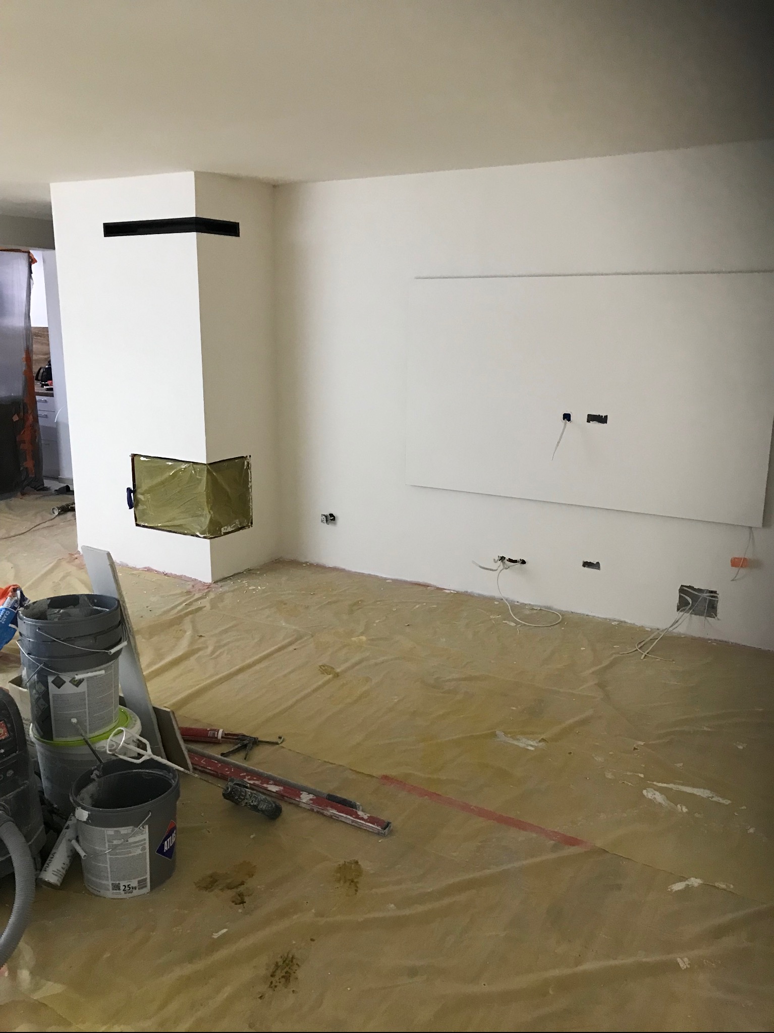 Wnętrze w trakcie wykończenia: białe ściany, zabudowa kominka z czarnym akcentem, przygotowane podłoże, widoczne narzędzia i materiały malarskie. Etap prac wykończeniowych.