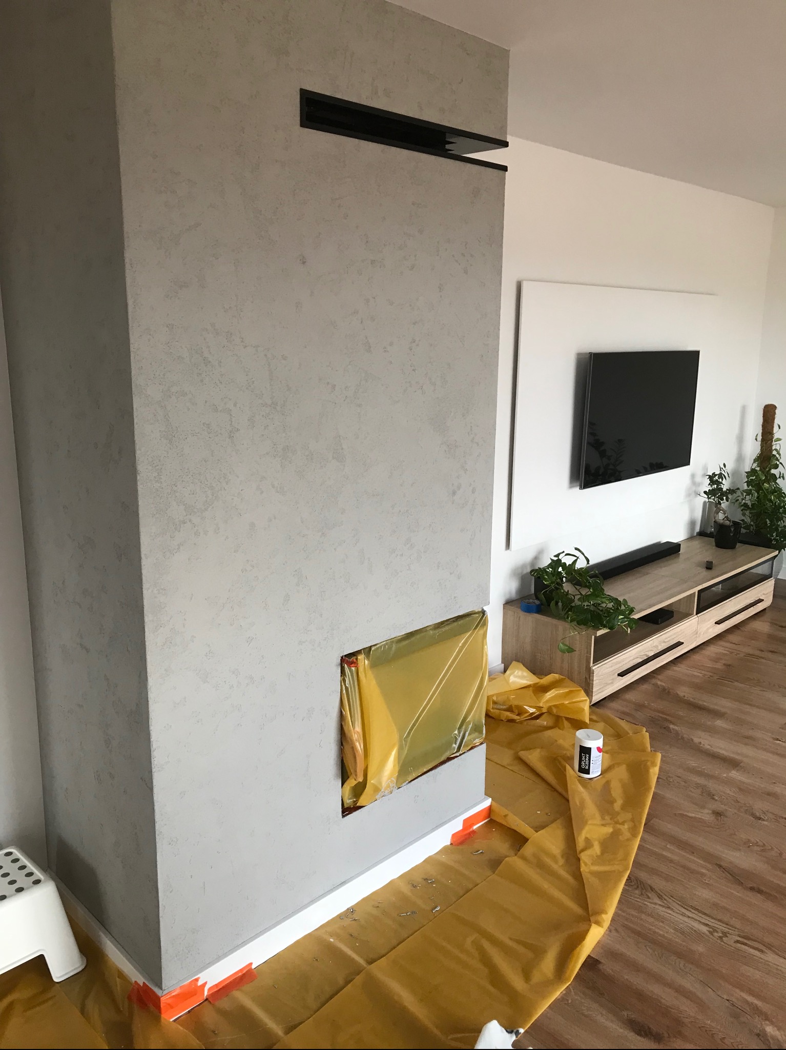 Ściana z efektem dekoracyjnym w odcieniach szarości, zabezpieczona folią malarską. Widoczny fragment salonu z telewizorem i roślinami. Etap prac wykończeniowych.