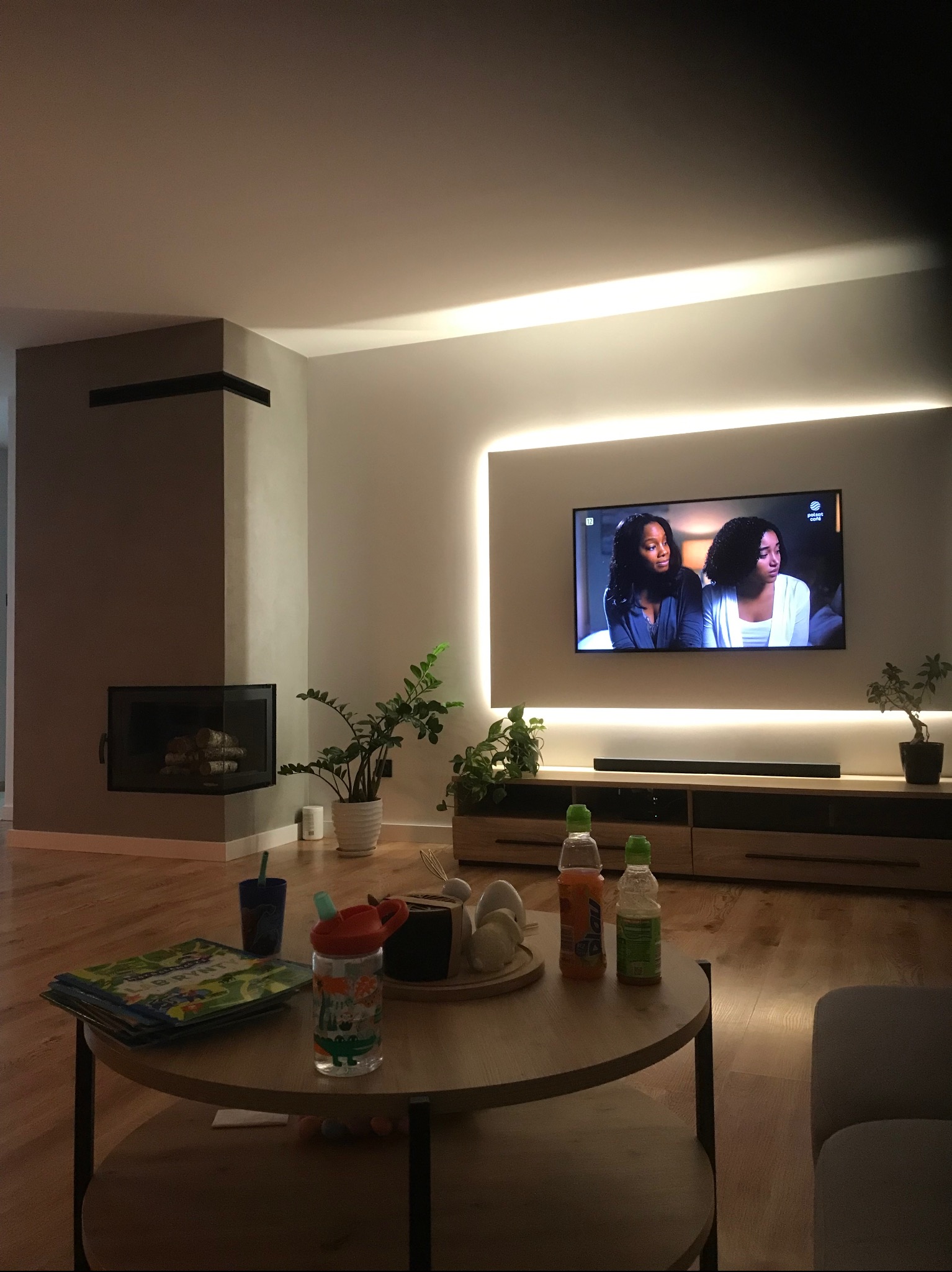 Nowoczesny salon z kominkiem i podświetlanym telewizorem. Minimalistyczny design, drewniana podłoga, rośliny doniczkowe. Ciepłe oświetlenie tworzy przytulną atmosferę.