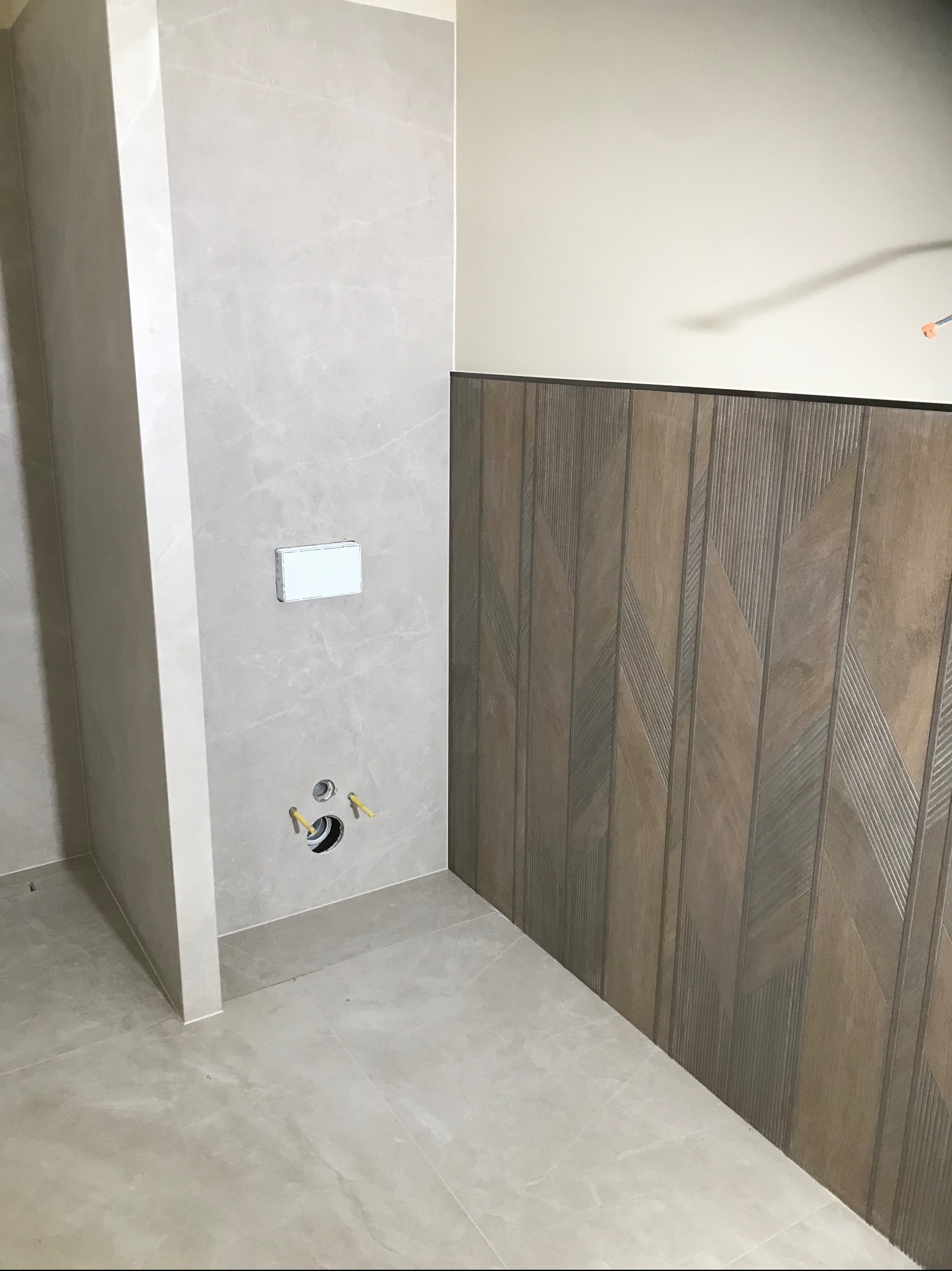 Wykończona łazienka z płytkami imitującymi marmur i drewno ułożonymi w jodełkę. Widoczne przyłącza do toalety podtynkowej i fragment podłogi z płytek.