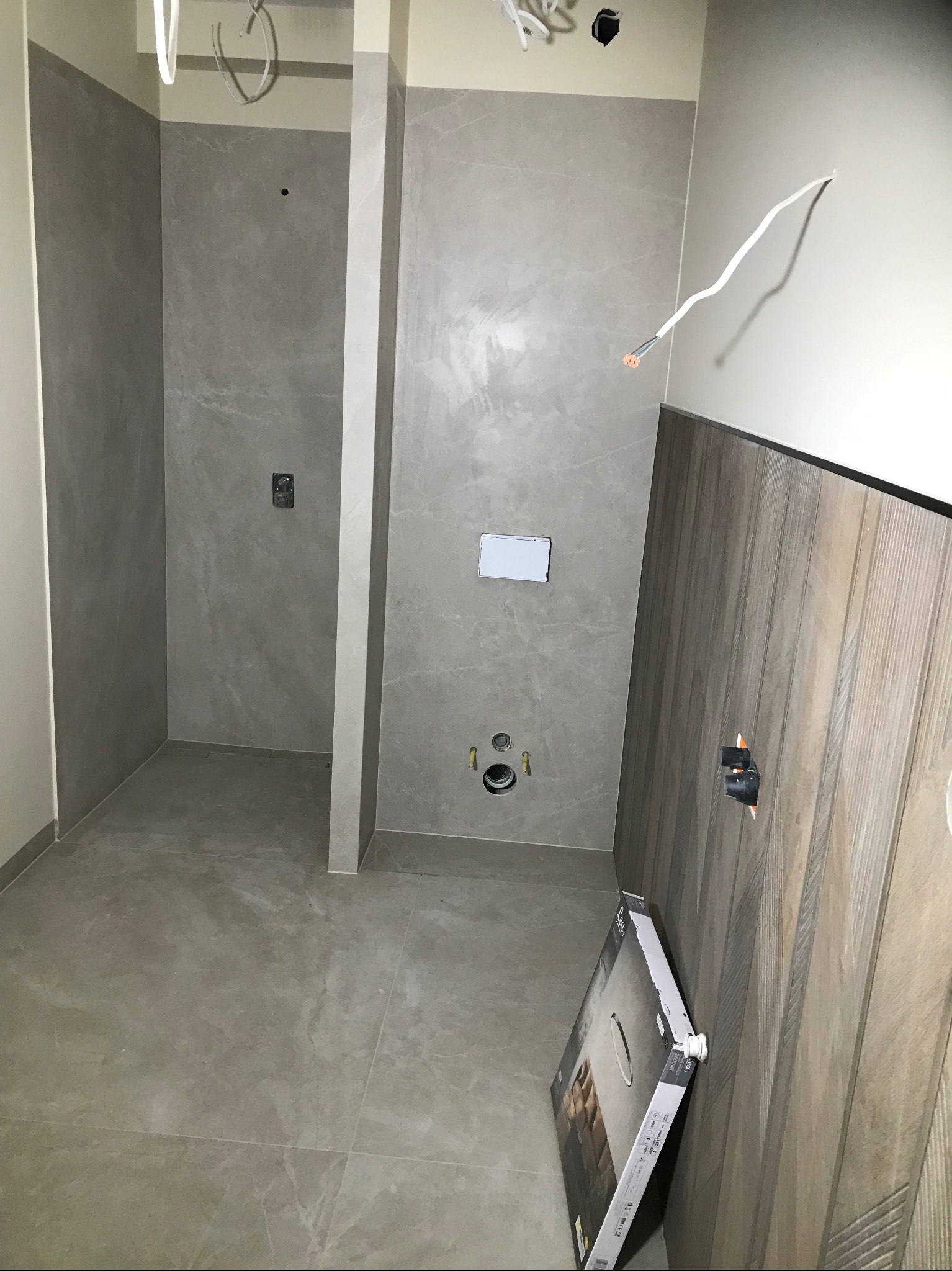 Wnętrze łazienki w trakcie wykańczania: szare płytki na ścianach i podłodze, częściowo wykończona drewniana boazeria, widoczne instalacje i karton z elementem wyposażenia.