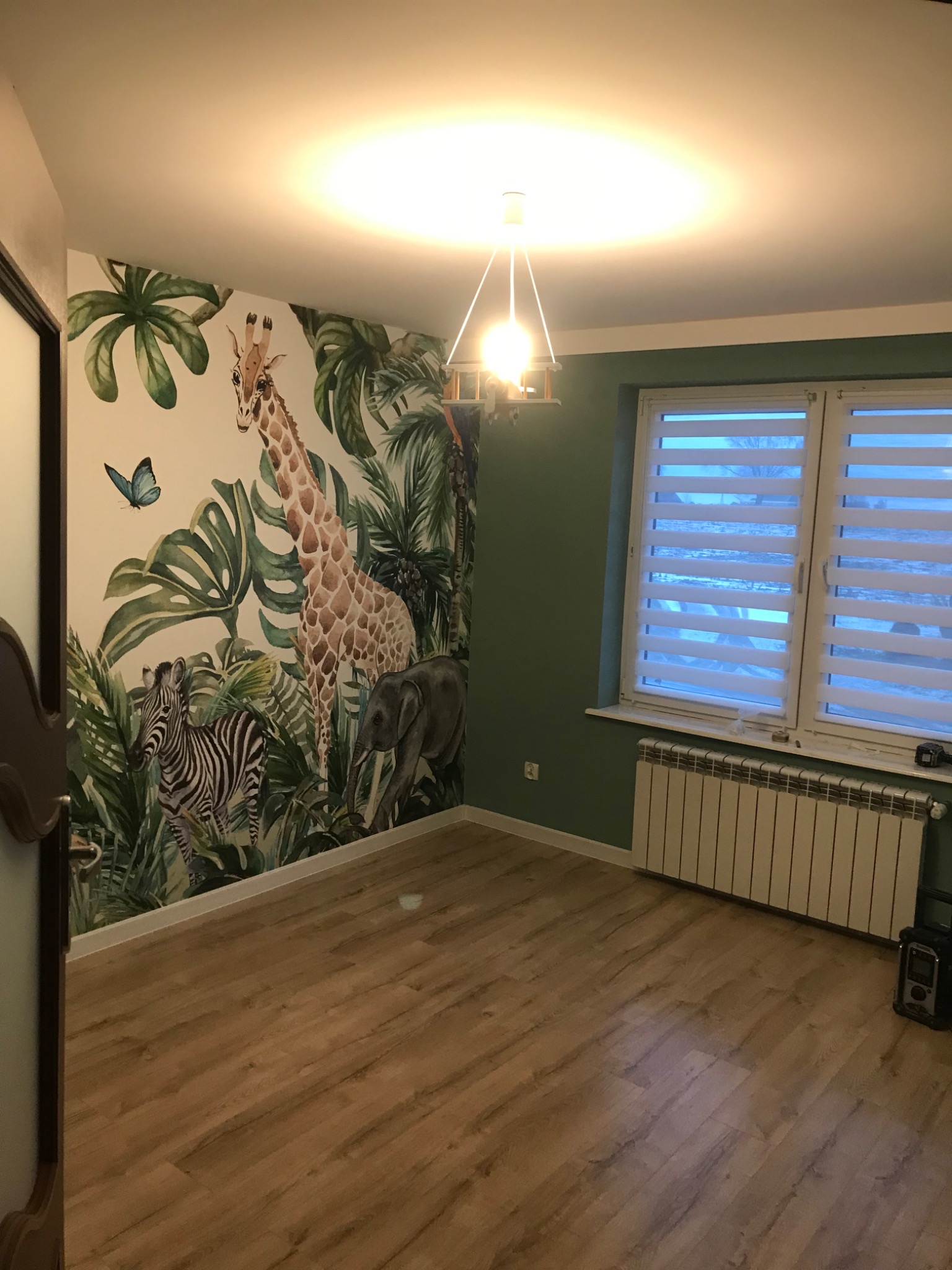 Pokój dziecięcy po remoncie: fototapeta z motywem dżungli (zebra, żyrafa, słoń), drewniana podłoga, okno z żaluzjami, zielona ściana. Nowoczesne oświetlenie.