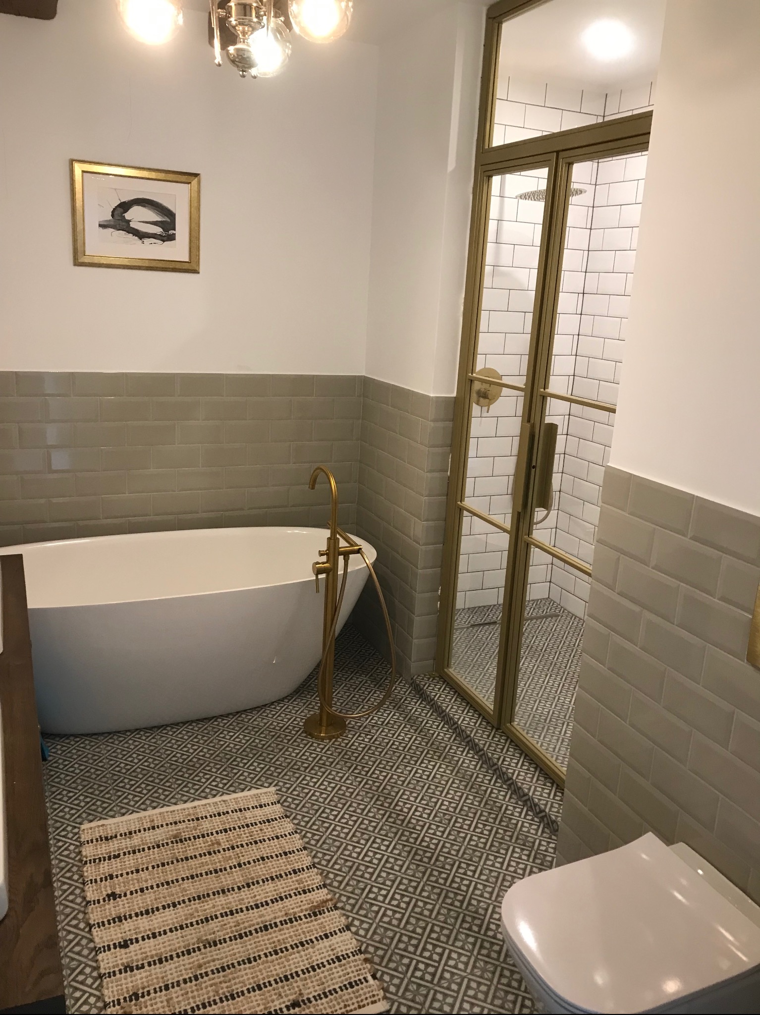 Elegancka łazienka z wolnostojącą wanną, złotą armaturą i kabiną prysznicową w stylu loft, wykończona szarą płytką i wzorzystą podłogą. Nad wanną abstrakcyjny obraz w złotej ramie.