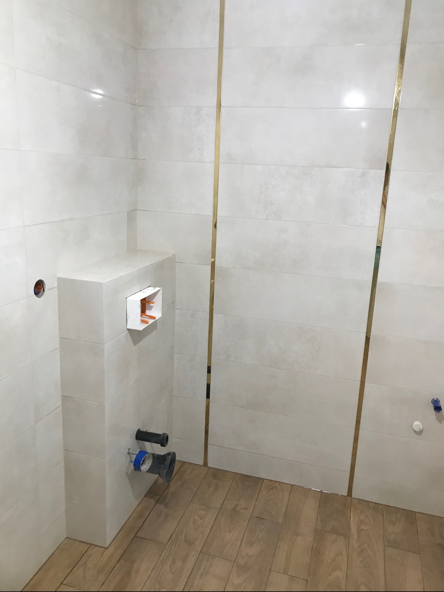 Białe, poziome płytki z wąskimi złotymi listwami w łazience, zabudowa stelaża WC z widocznymi przyłączami kanalizacyjnymi i drewnopodobną podłogą.