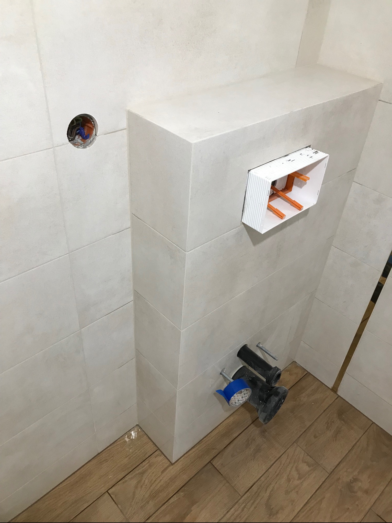 Ścianka z zabudową WC w trakcie montażu, widoczne przyłącza kanalizacyjne i instalacyjne, płytki ceramiczne na ścianach i drewnopodobne panele na podłodze.