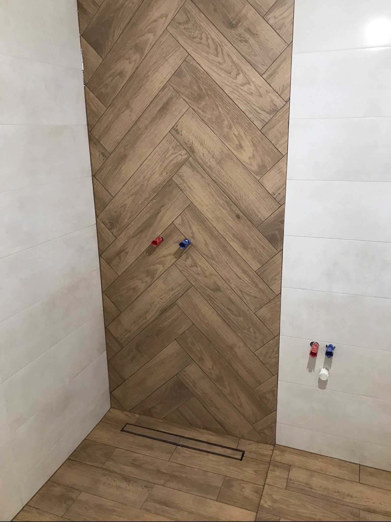 Wykończona łazienka z drewnopodobnymi płytkami ułożonymi w jodełkę na ścianie i podłodze, kontrastującymi z białymi płytkami na sąsiedniej ścianie. Widoczne przyłącza wodne.