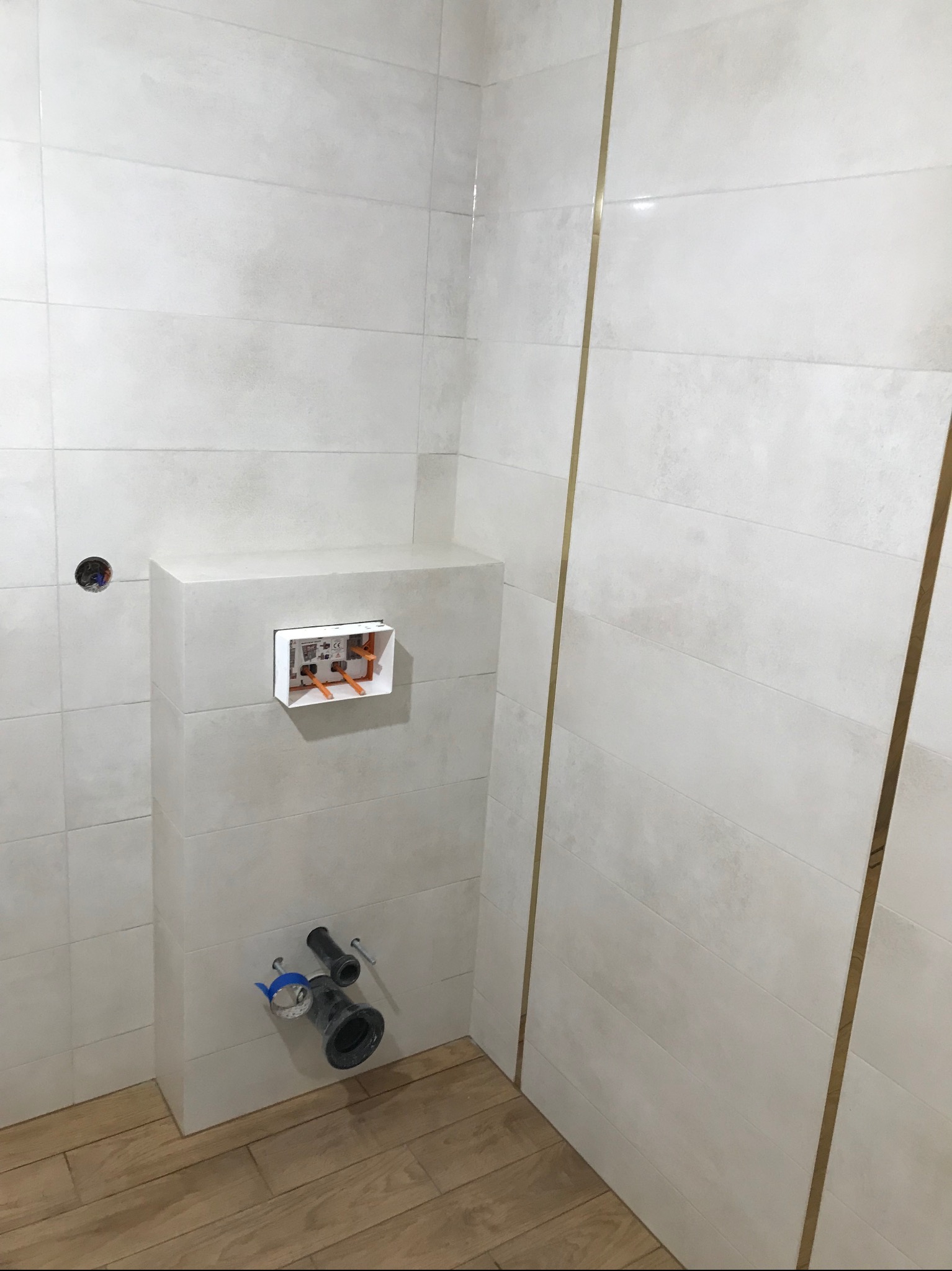 Biała, wykafelkowana łazienka w trakcie montażu podtynkowego stelaża WC. Widoczne przyłącza kanalizacyjne i instalacja. Minimalistyczny design z akcentami w kolorze złota.