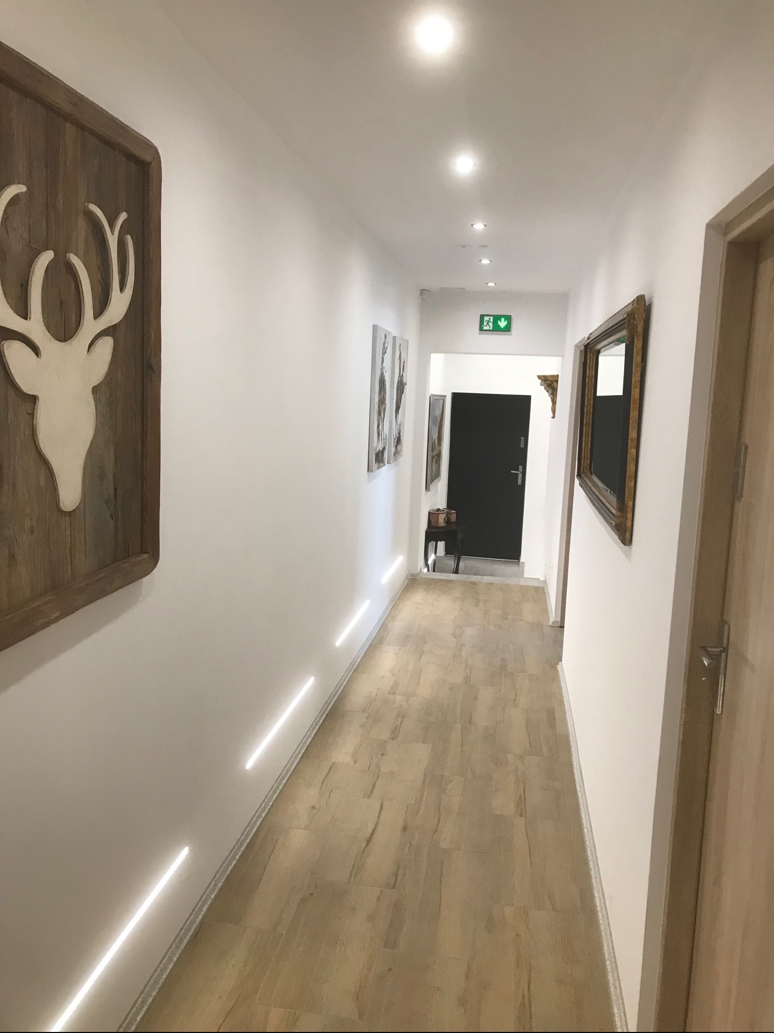 Korytarz z drewnianą podłogą, oświetleniem LED w listwach przypodłogowych i dekoracjami ściennymi, w tym rzeźbą głowy jelenia w drewnianej ramie.