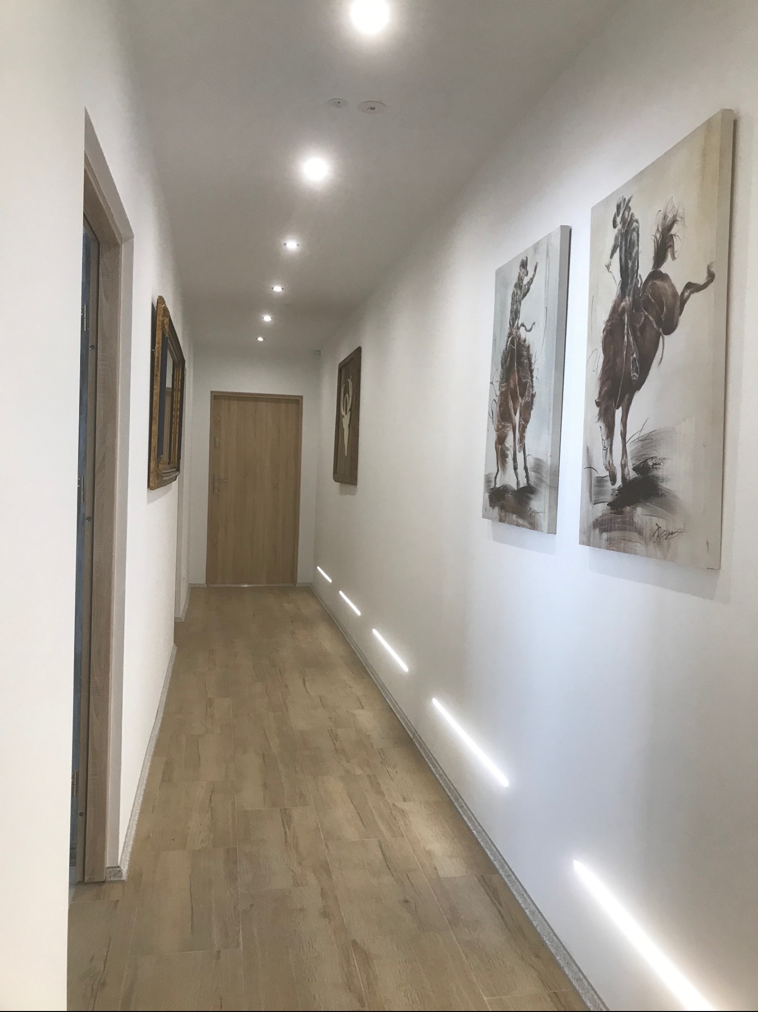 Długi korytarz z drewnianą podłogą, oświetleniem podłogowym i sufitowym. Na ścianach wiszą obrazy z motywem koni. Widoczne drzwi i ramy obrazów.