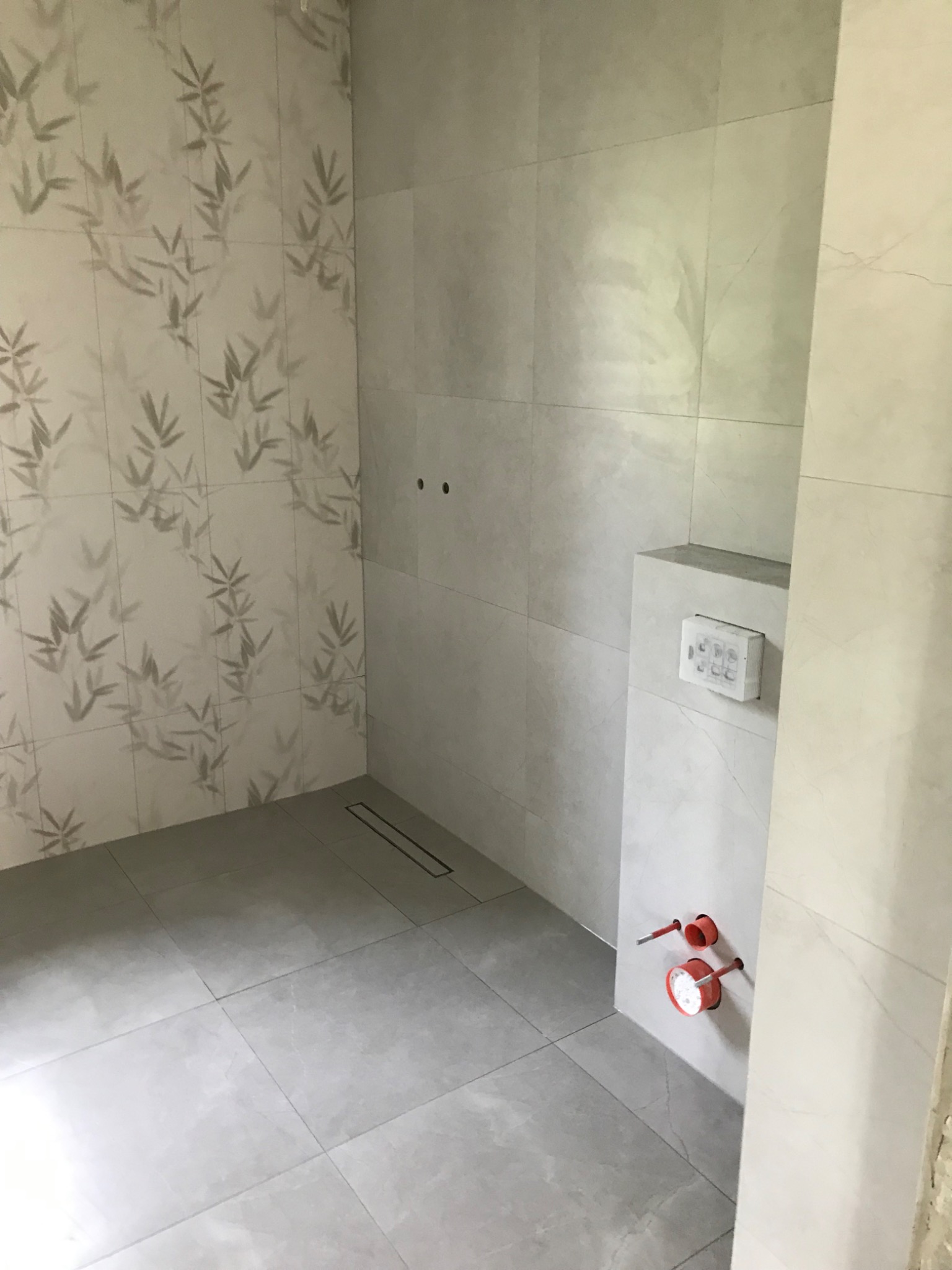 Wykończona łazienka z szarymi płytkami, wzorem bambusa na ścianie, odpływem liniowym i instalacją wodną. Minimalistyczny design, nowoczesne wykończenie.